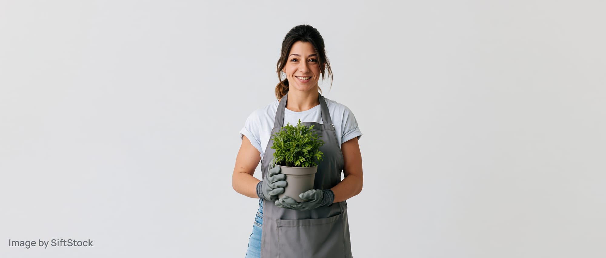 Woman in grey apron holding potted green plant, smiling, studio portrait