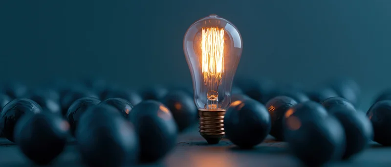 Single glowing edison filament lightbulb standing upright among unlit bulbs on dark surface