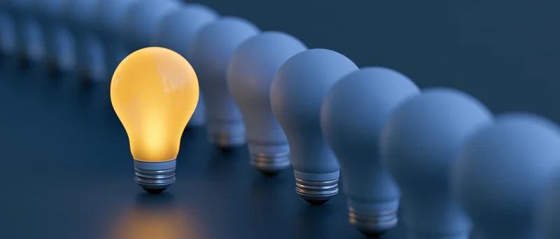 Glowing yellow lightbulb standing in a row of unlit bulbs on a dark surface