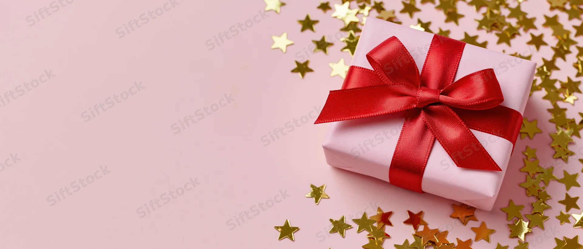 Pink gift box with red satin ribbon bow surrounded by gold star confetti on a pink surface