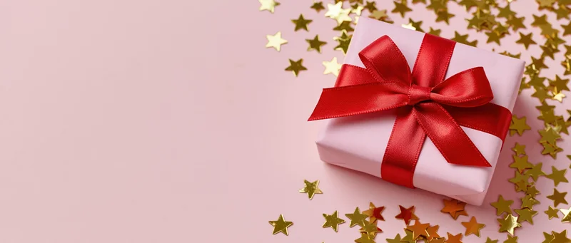 Pink gift box with red satin ribbon bow surrounded by gold star confetti on a pink surface