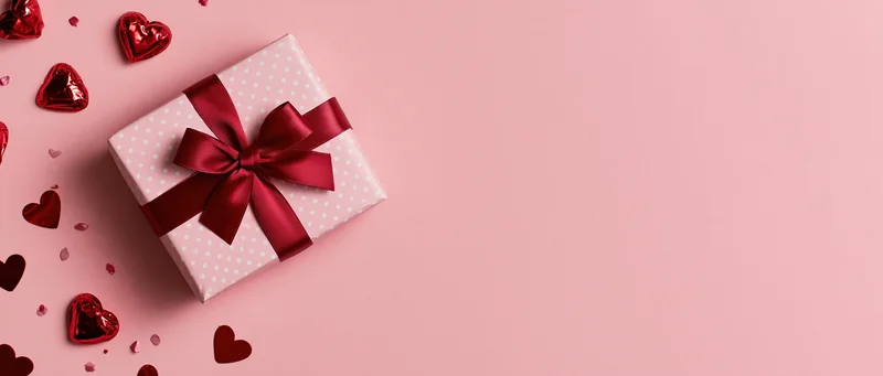 Gift box wrapped in pink polka-dot paper with red ribbon and bow, surrounded by red hearts on pink background