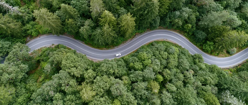 Aerial view of a winding two-lane road through dense green forest with a white car driving along it