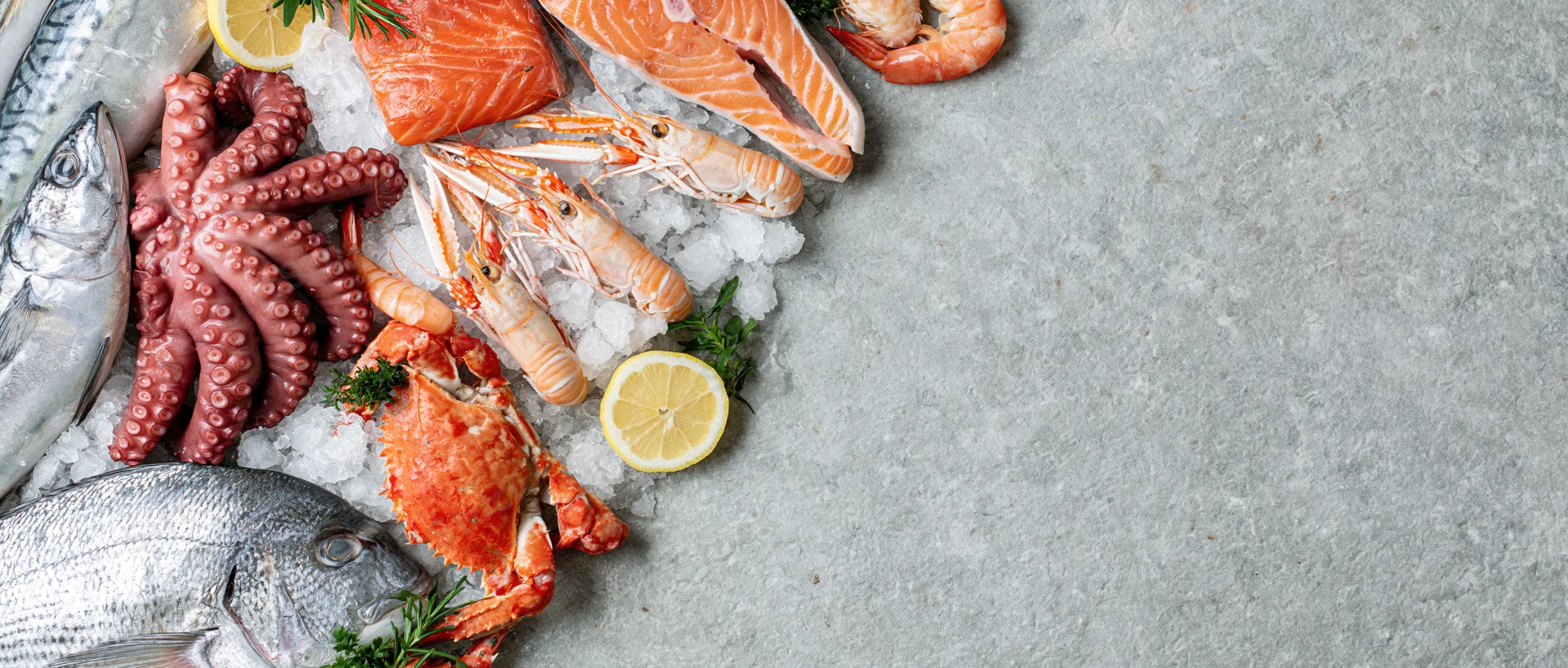 Fresh raw seafood including salmon, shrimp, octopus and crab arranged on crushed ice on a gray stone surface with lemon and herbs
