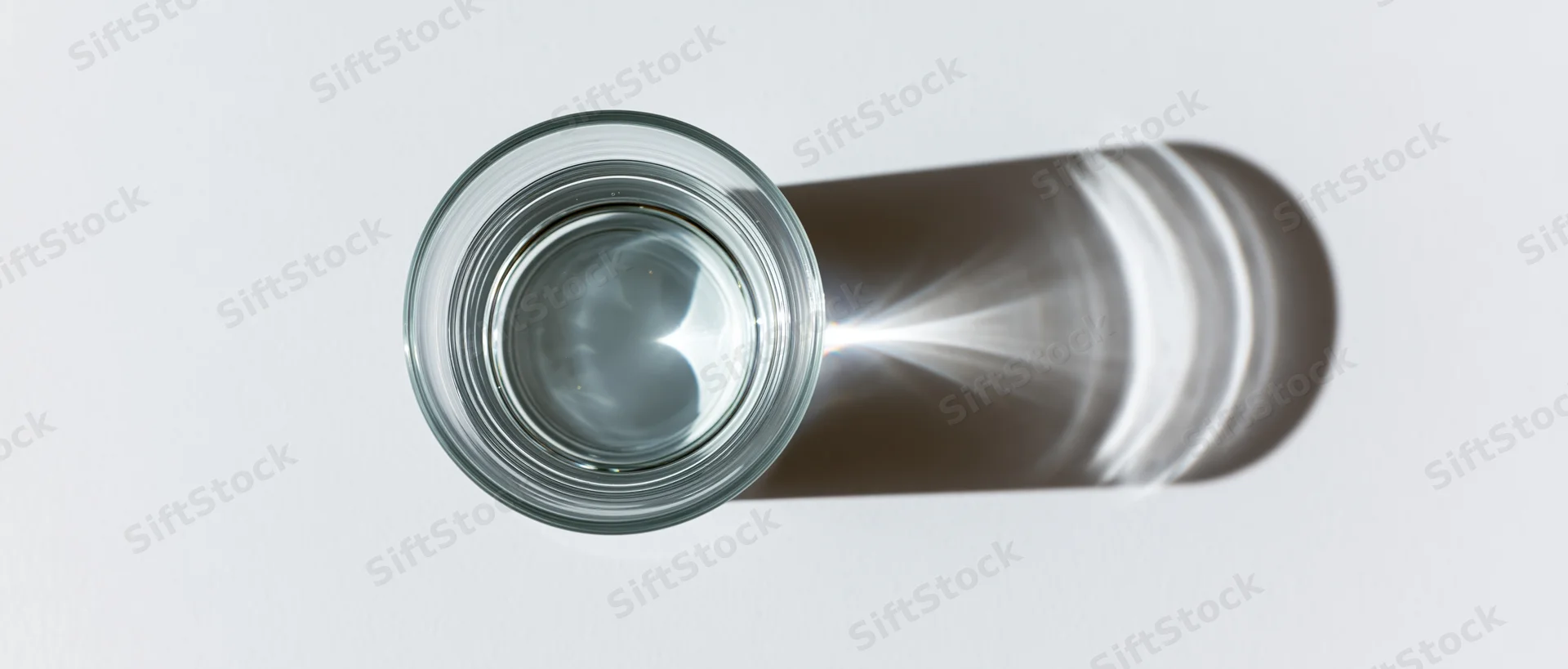 Overhead view of a clear glass of water on a white surface casting a shadow with light refraction visible