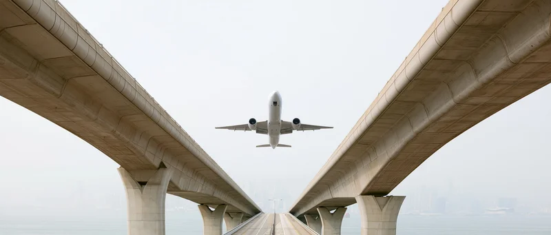 Commercial passenger jet flying low between two diverging concrete bridge spans over water with city skyline in distance
