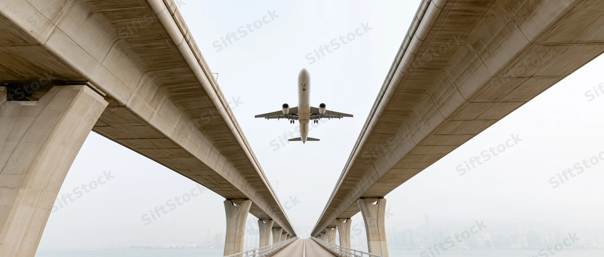 Commercial airplane flying low between concrete bridge supports over water toward city skyline