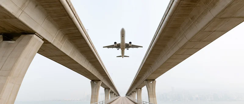 Commercial airplane flying low between concrete bridge supports over water toward city skyline