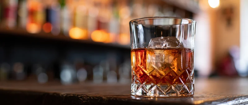 Amber whiskey in a faceted crystal rocks glass with ice on a wooden bar counter, warm ambient lighting in background