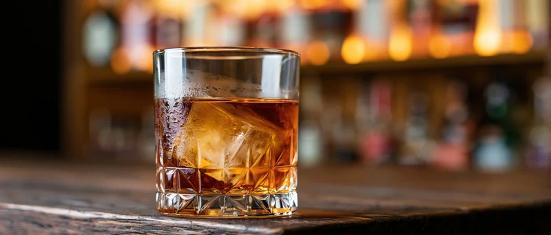Amber whiskey with ice in a crystal rocks glass on a wooden bar counter