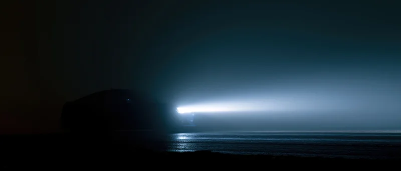 Dark silhouette of a sports car on an open road at night with headlights glowing over water