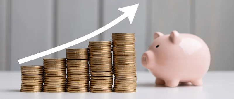 Stacks of gold coins increasing in height with a white arrow pointing upward beside a pink piggy bank on a white surface