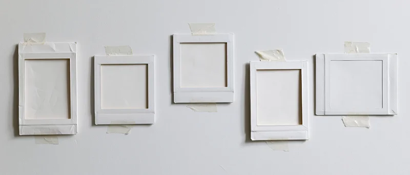 Five blank white instant photo frames taped in a row on a plain white wall