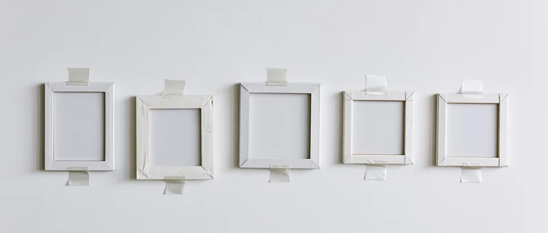 Five blank white photo frames taped to a plain white wall in a row