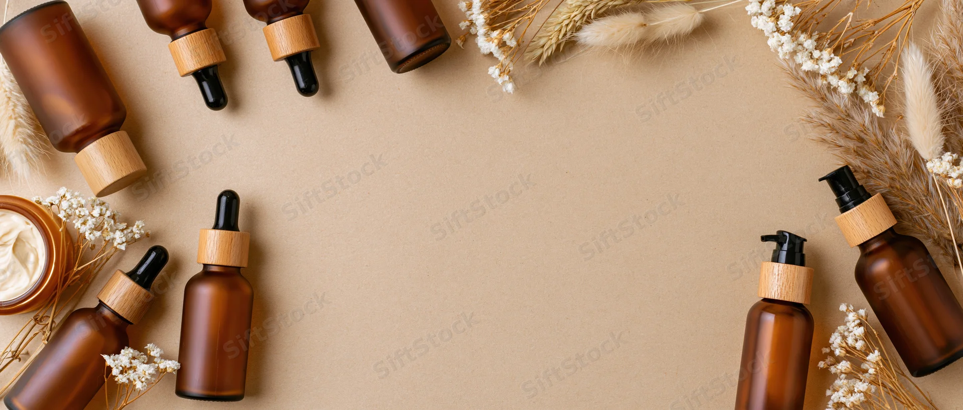 Amber glass skincare bottles with wooden caps and dried flowers arranged in a flat lay on beige background
