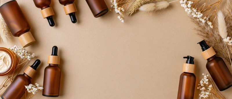 Amber glass skincare bottles with wooden caps and dried flowers arranged in a flat lay on beige background