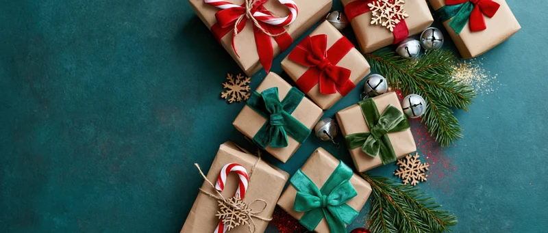 Kraft paper gift boxes with red and green bows, candy canes, snowflakes, pine sprigs, and jingle bells arranged on a dark teal surface in a flat lay composition with copyspace