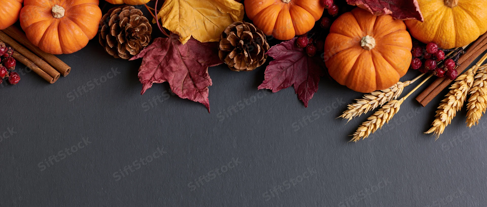 Small orange pumpkins and dried maple leaves arranged with pine cones, cinnamon sticks, berries, and wheat stalks along the top of a dark gray surface