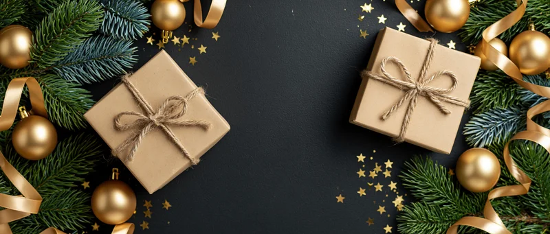 Two kraft paper gift boxes tied with twine bows arranged on dark surface with pine branches, gold ornaments and ribbons around edges