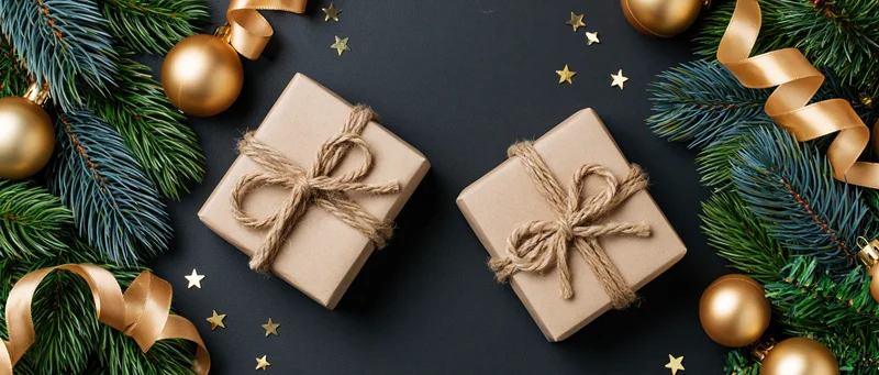 Two kraft paper gift boxes tied with twine bows on dark surface surrounded by pine branches, gold ornaments and stars