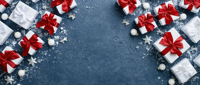 White gift boxes with red satin bows and silver ornaments arranged on blue surface with snow, forming border with central copyspace