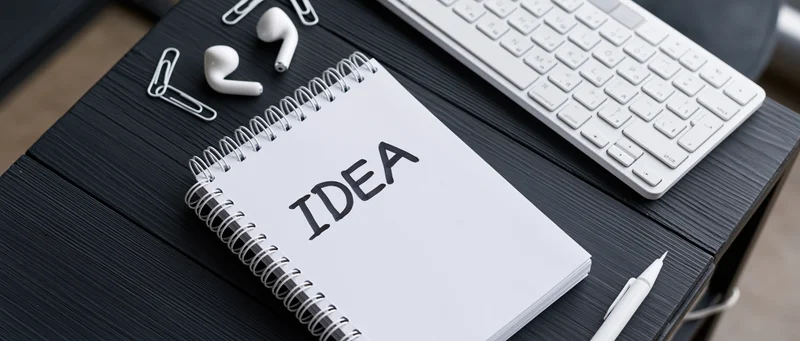 Notebook with idea written on it on a dark wooden desk with keyboard, earbuds, pen and paperclip