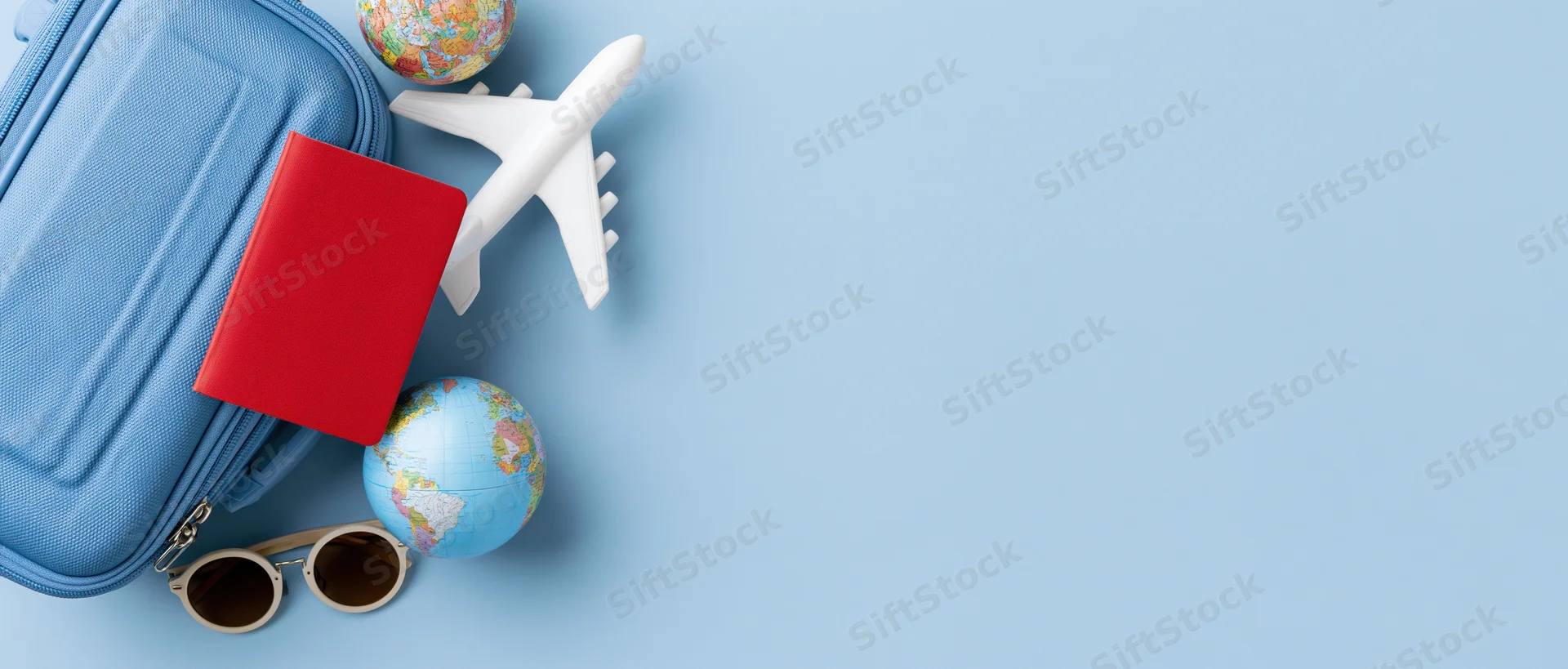 Travel items including suitcase, passport, airplane, globe and sunglasses arranged on blue surface with copyspace