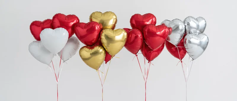 Four bunches of heart-shaped foil helium balloons in red, gold, silver and white arranged in a row against a plain background
