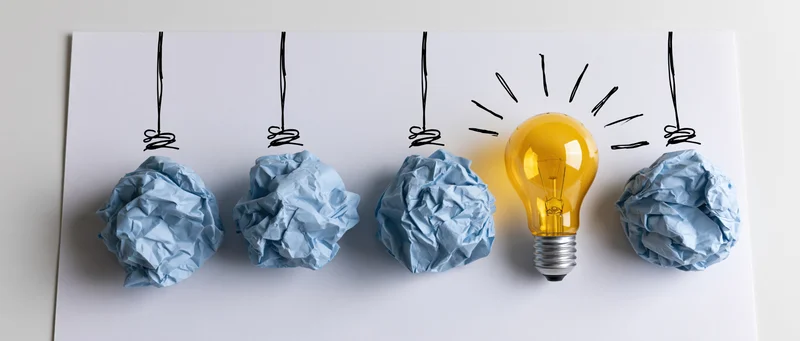 Lightbulb glowing among crumpled blue paper balls on white surface with hand-drawn strings and rays