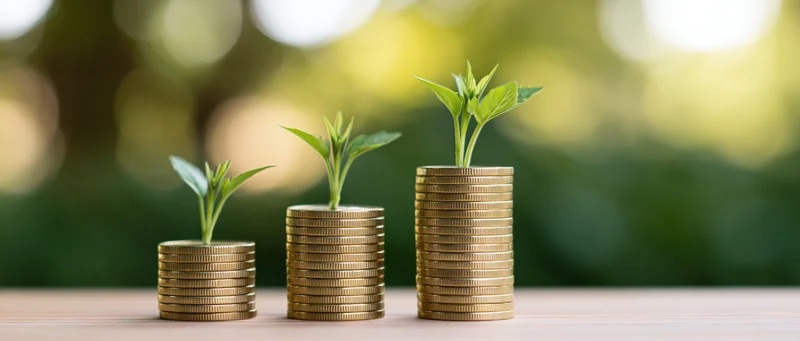 Three stacks of gold coins with growing plants on top arranged in ascending height on a wooden table outdoors