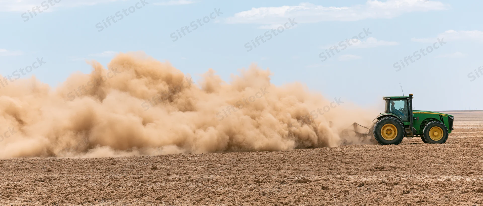Green john deere tractor pulling disc cultivator across dry field under blue sky