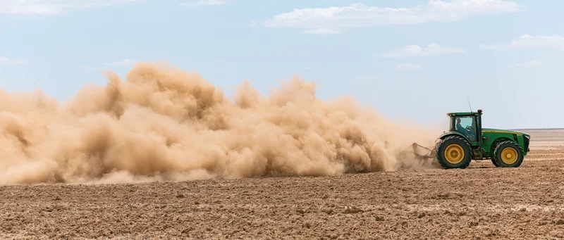 Green john deere tractor pulling disc cultivator across dry field under blue sky