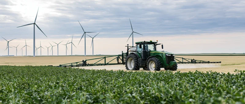 Green tractor pulling crop sprayer across flat farmland with wind turbines in background