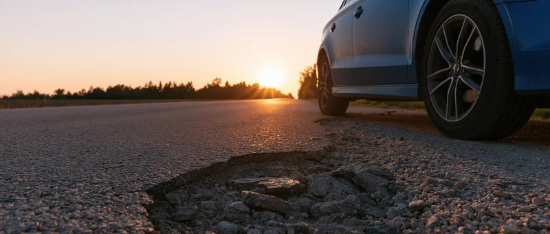 Closeup of a pothole on cracked asphalt road with a blue car parked nearby at sunset