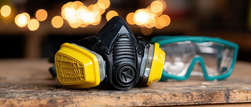 Half-face respirator with yellow filter cartridges and teal safety goggles resting on a wooden workbench