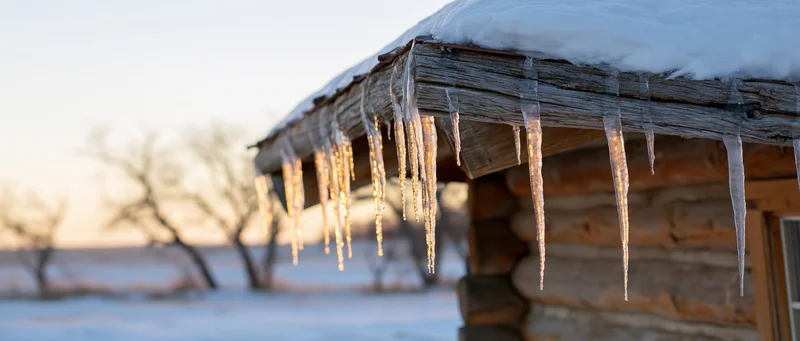 Icicles hanging from the eave of a rustic log cabin in a snowy rural landscape at sunrise