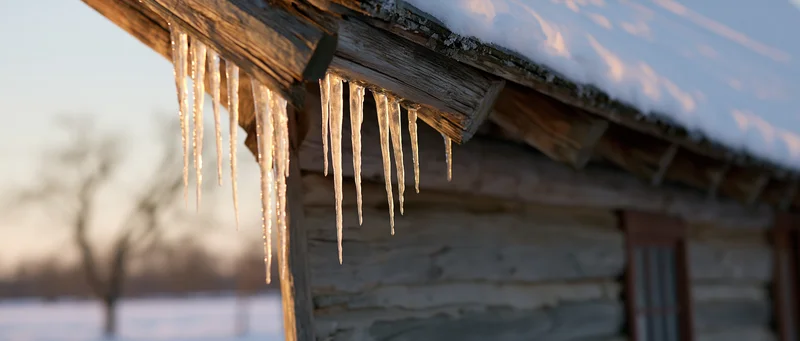 Icicles hanging from the eave of a rustic log cabin in a snowy rural landscape at twilight