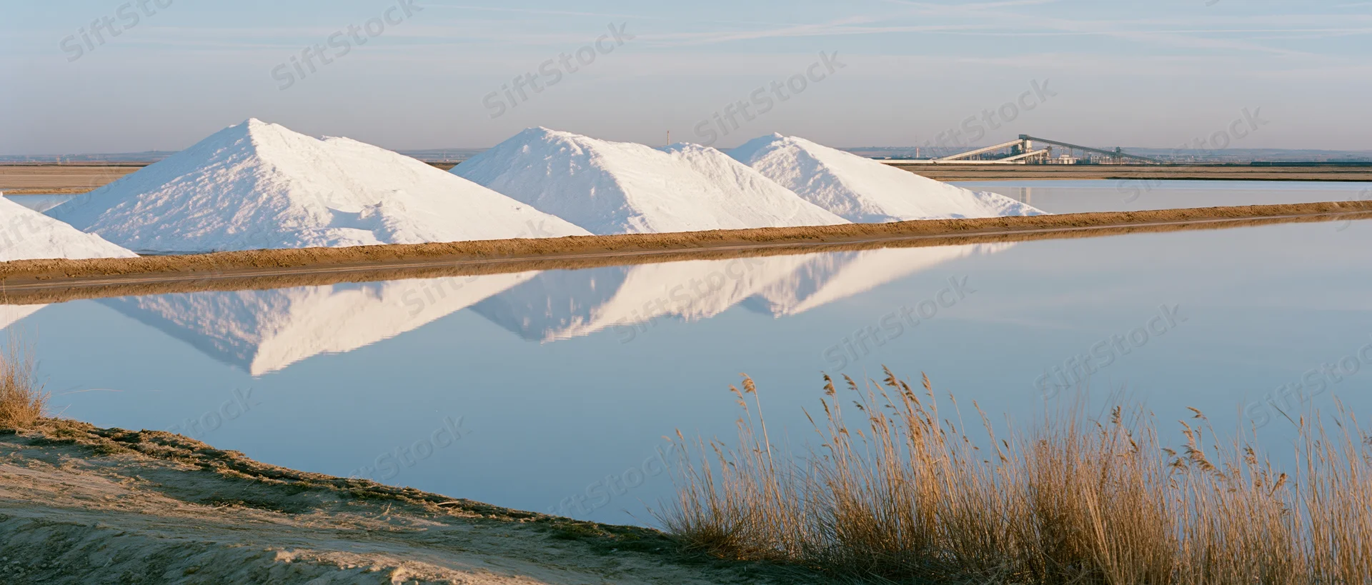 Large white salt mounds piled at an industrial salt flat with calm water reflecting them under a clear sky