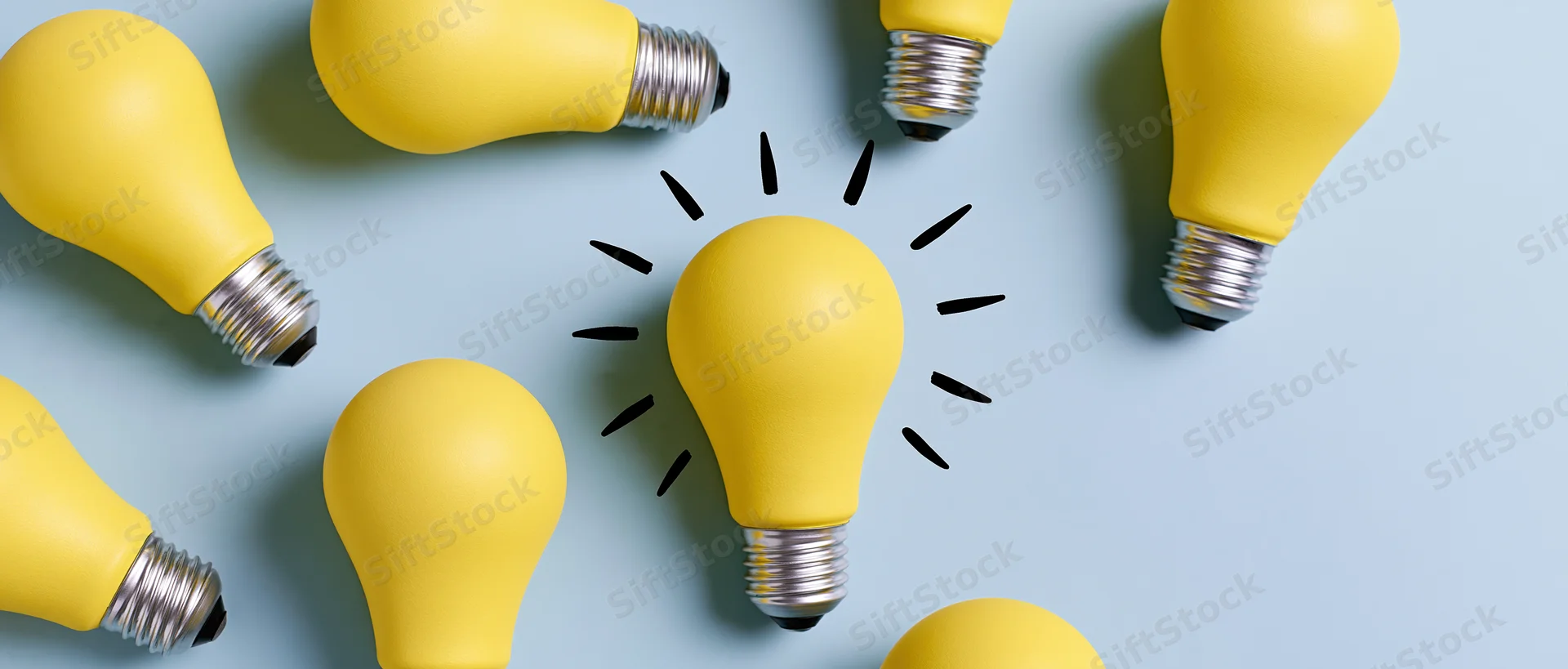 Multiple matte yellow light bulbs scattered on a light blue surface with one centered and glowing with radiating lines