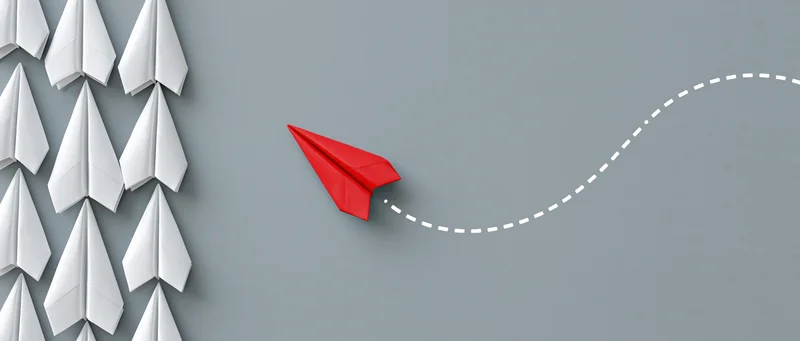 Red paper airplane breaking away from a group of white ones on gray background