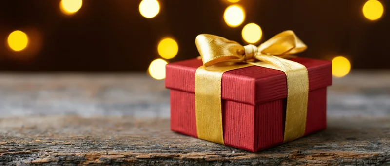 Red gift box with gold satin ribbon and bow on rustic wooden table against warm bokeh lights