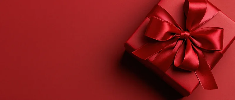 Red gift box with large satin bow on red surface, positioned in corner with copyspace