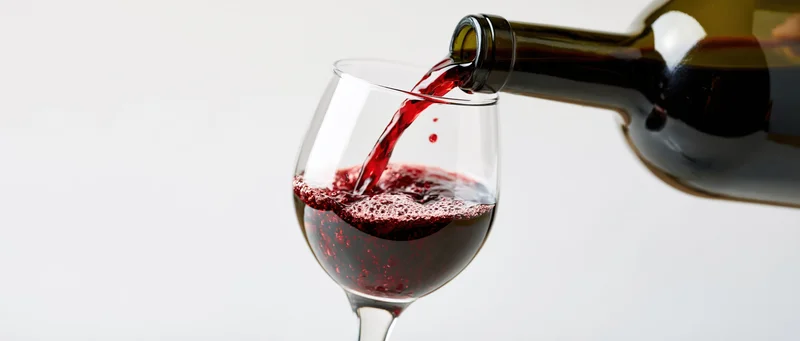Red wine pouring from a dark green bottle into a clear glass on white background