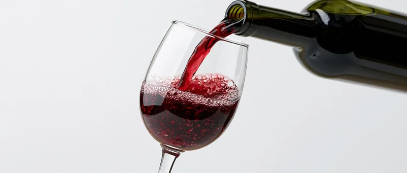 Red wine pouring from a dark green bottle into a clear wine glass against white background