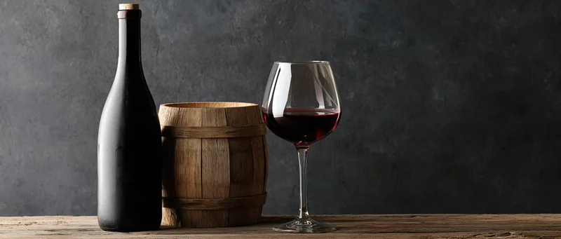 Red wine bottle and half-filled glass beside a small wooden barrel on a worn wooden table against a dark textured background