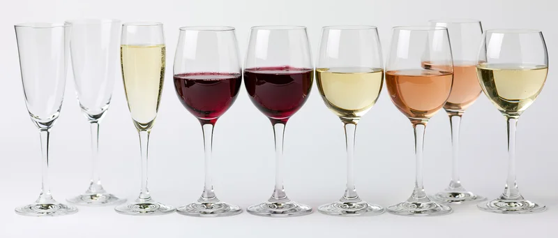 Row of wine and champagne glasses filled with different colored liquids against white background