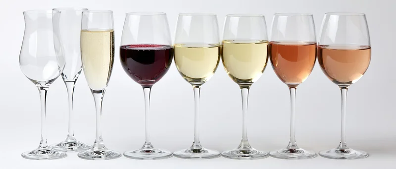 Seven wine and champagne glasses in a row filled with different colored wines against a white background