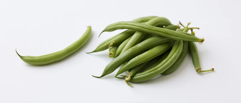 Single fresh green bean lying flat beside a small loose pile on white background
