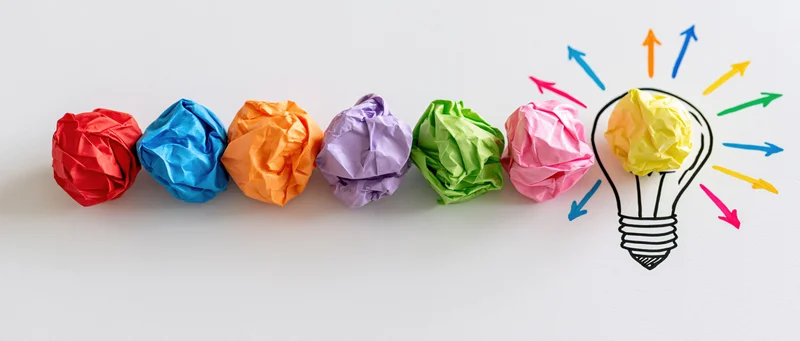 Row of colorful crumpled paper balls leading to a hand-drawn lightbulb with arrows radiating outward on white background