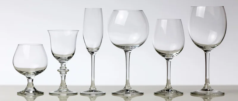 Six empty glassware pieces arranged in a row from shortest to tallest on a reflective surface against a white background